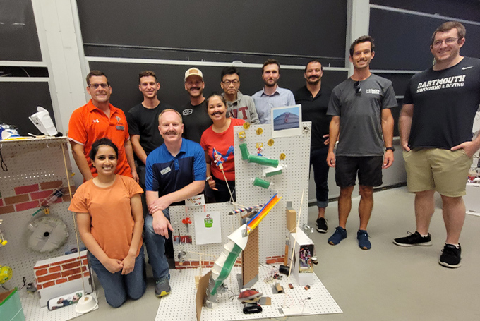 A group of students gather around a complicated homemade Rube Goldberg device.