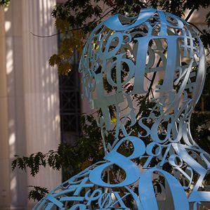 The Alchemist, a sculpture made up of large curved numbers and symbols in the shape of the head and shoulders of a person, is in the foreground. In the background are the pillars and steps of the main entrance to MIT.