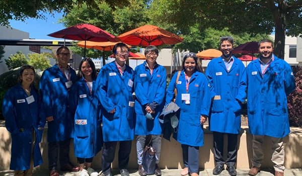 A group of students in long blue lab coats, standing outside in partial sun and partial shadow.