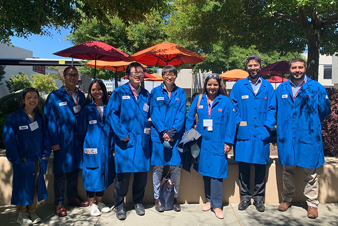 A group of students in long blue lab coats, standing outside in partial sun and partial shadow.