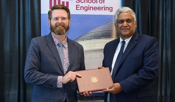 Steven Derocher, a tall white man with red hair, a beard, and glasses, receives an award from Anantha Chandrakasan, a slightly shorter man with brown skin and white hair. They are standing in front of a banner for the MIT School of Engineering.