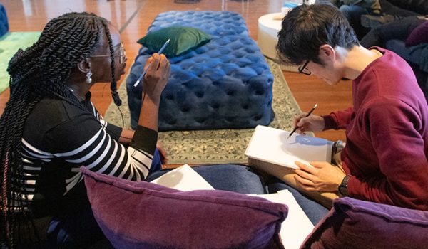 Two students sit on a couch together, with one explaining something and gesturing and the other drawing on a piece of paper.