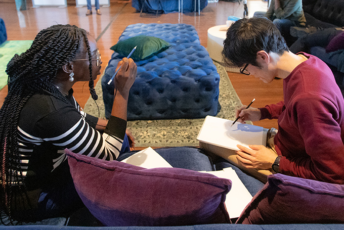 Two students sit on a couch together, with one explaining something and gesturing and the other drawing on a piece of paper.