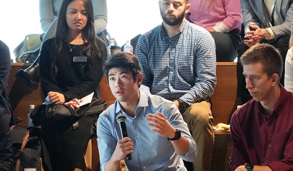 A group of students sitting on wooden risers. One holds a microphone and is asking a question, gesturing with his free hand.