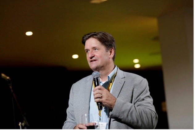 Bryan Moser stands with a microphone in hand, brightly lit against a dark ballroom background.