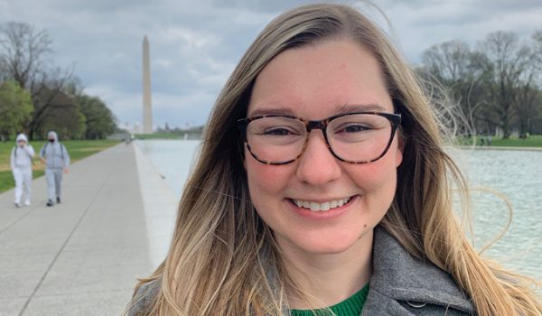 Morgan Orlovskiy, a woman with long blonde hair and glasses, smiles brightly at the camera. In the distance behind her is the Washington Monument, a tall obelisk.