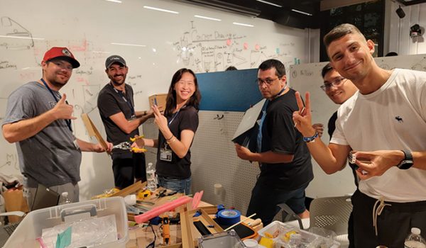 A group of students around a table scattered with tools. They're all smiling at the camera as they work on a project.