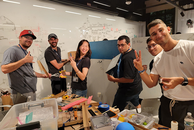 A group of students around a table scattered with tools. They're all smiling at the camera as they work on a project.